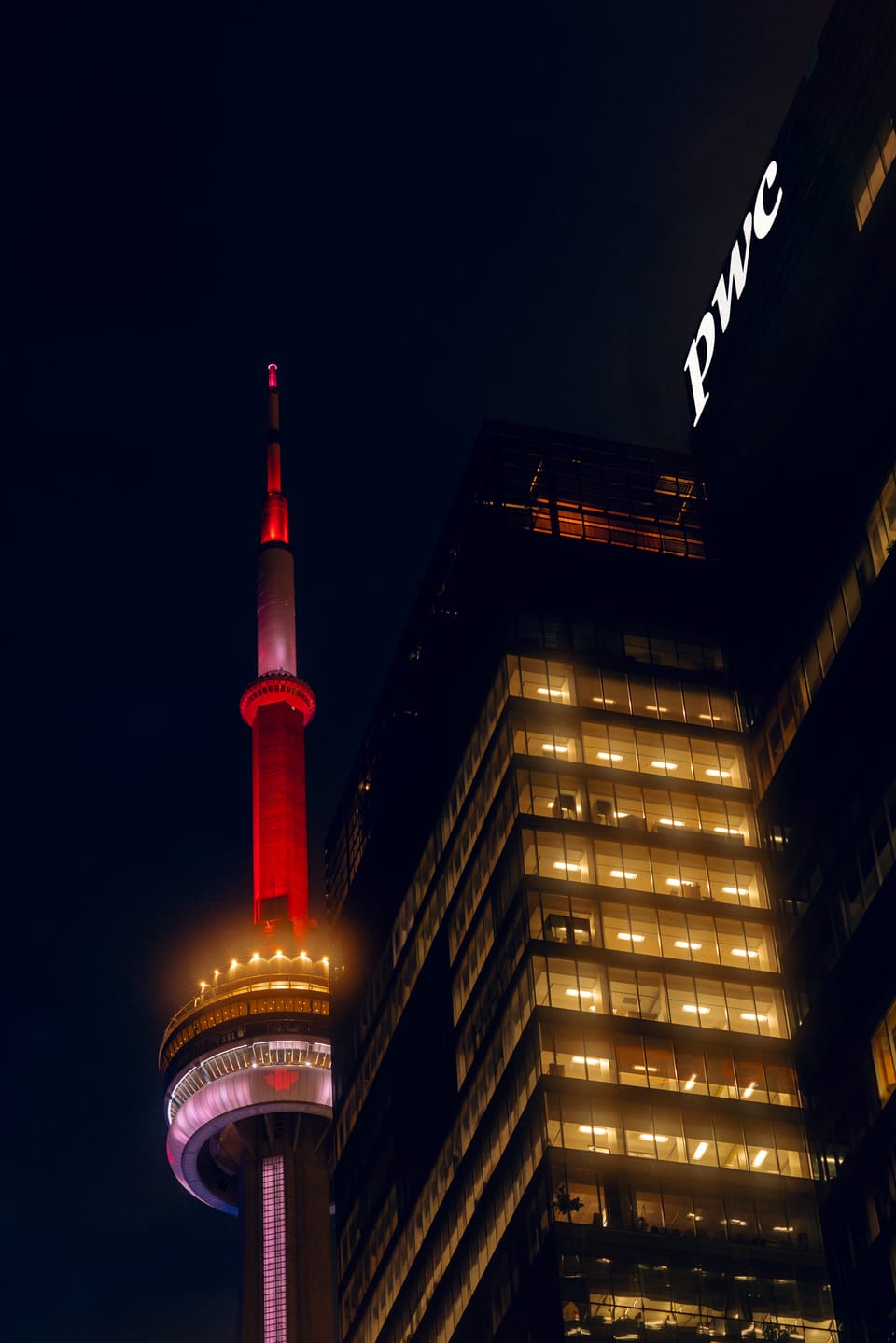The CN Tower glowing red at night beside a golden office tower in Toronto.