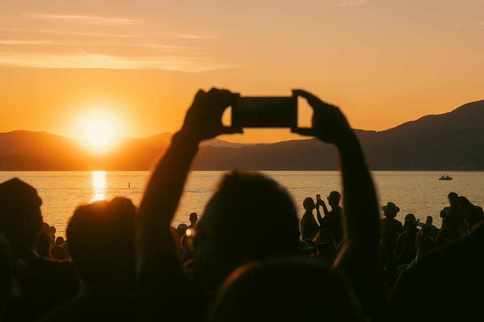 Silhouetted hands raise a phone to photograph a burning sunset over the bay.