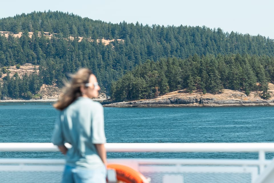 A woman in a pale blue shirt gazing from a ferry deck toward a forested coastline.