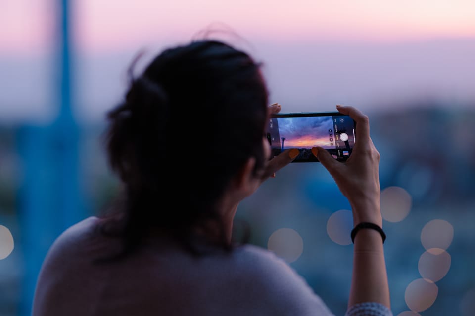 A woman seen from behind photographing a hazy city dusk on her phone.