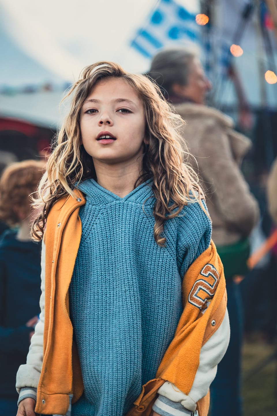 A young girl in a varsity jacket standing before a bright fairground.