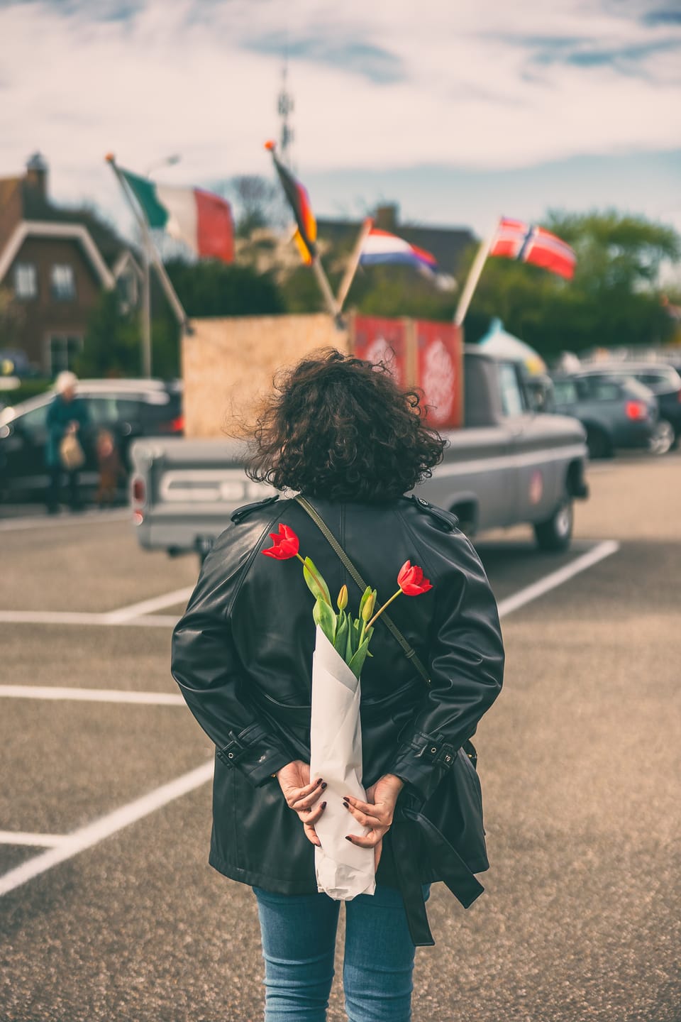 A woman in a leather jacket carrying tulips hidden behind her back in a parking lot.