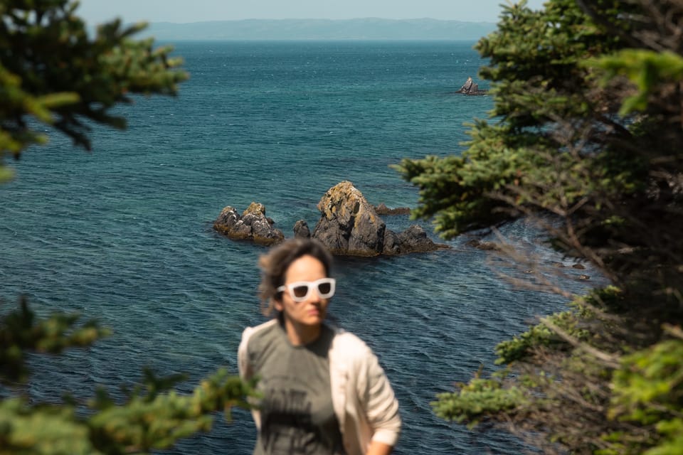 A woman in white sunglasses walking past sea rocks and evergreens on the coast.